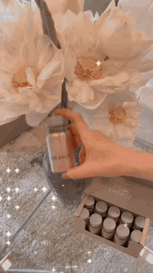 a person holding a bottle of emese next to a vase of white flowers