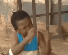 a man in a blue tank top is covering his mouth with his hand while sitting in front of a window .